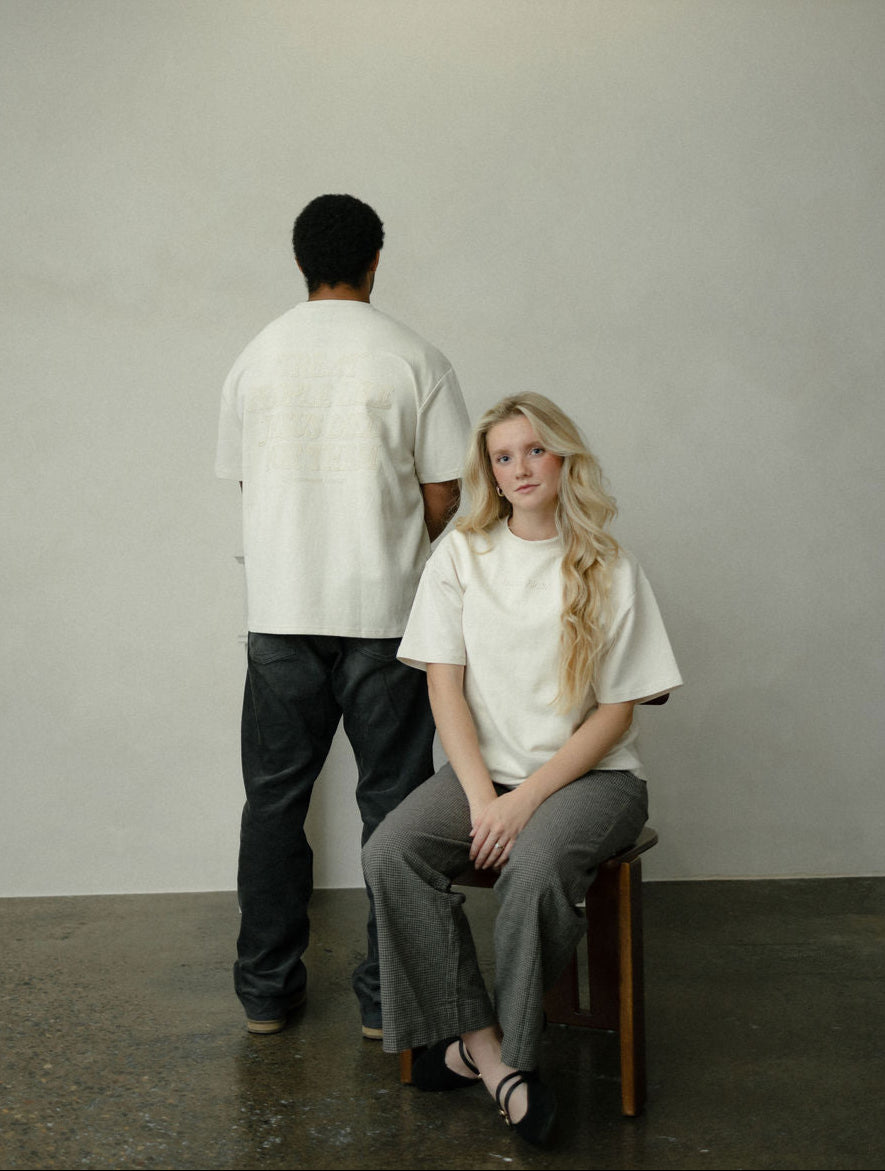 Two people, one standing and one sitting on a stool, wearing white tops against a plain background.